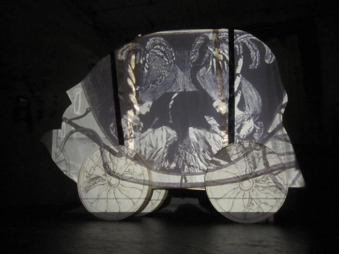 Kate Ruddle's Marie Antoinette Carriage piece that is 5 feet tall by 6 feet wide and made of silk fabric stretched over bend wood. The Carriage has a still image projected on it of a cartoon from Marie Antoinette's time . The Cartoon depicts two women quietly reading inside a carriage with embellished tall hair and extravagent feathers that fill the carriage space to the brim.